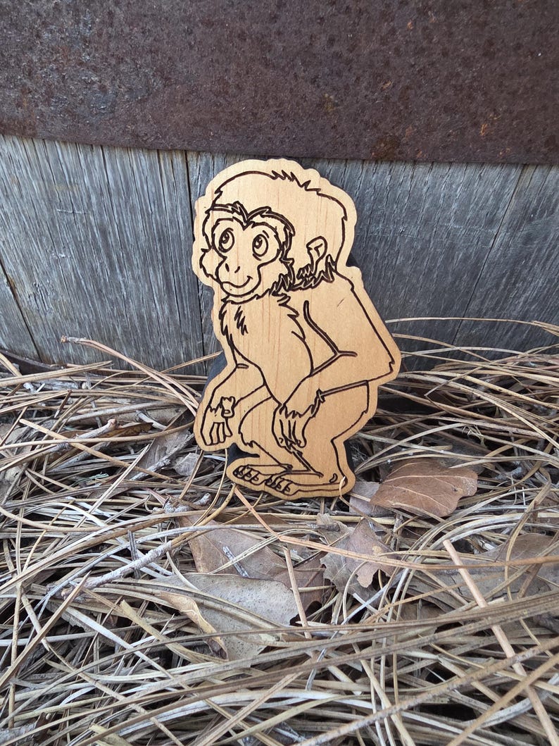 May include: A light-colored wooden cutout shaped like a monkey, featuring dark laser-engraved outlines that define the facial features, fur texture, and body posture of the primate. The figure is depicted in a crouching position with a gentle expression.