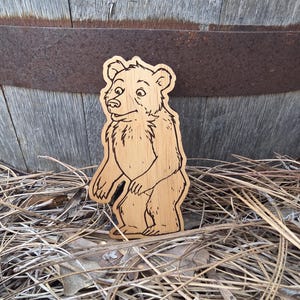 May include: A light brown wooden cutout shaped like a standing bear with dark brown laser-engraved outlines detailing the fur, facial features, and paws. The bear is depicted in a side profile view with its head turned slightly toward the front.