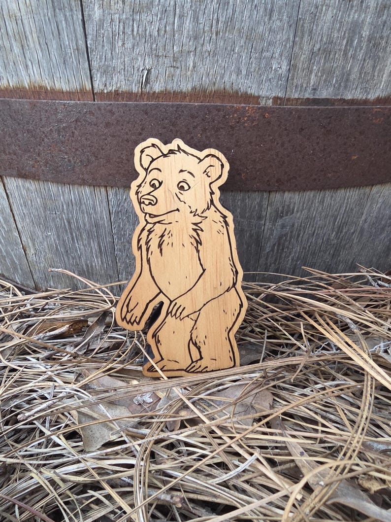 May include: A light brown wooden cutout shaped like a standing bear with dark brown laser-engraved outlines detailing the fur, facial features, and paws. The bear is depicted in a side profile view with its head turned slightly toward the front.