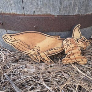 May include: A light brown wooden cutout featuring a detailed engraving of a humpback whale swimming alongside a mermaid with long flowing hair. The wood grain is visible throughout the surface, and the edges of the shapes are dark and defined.