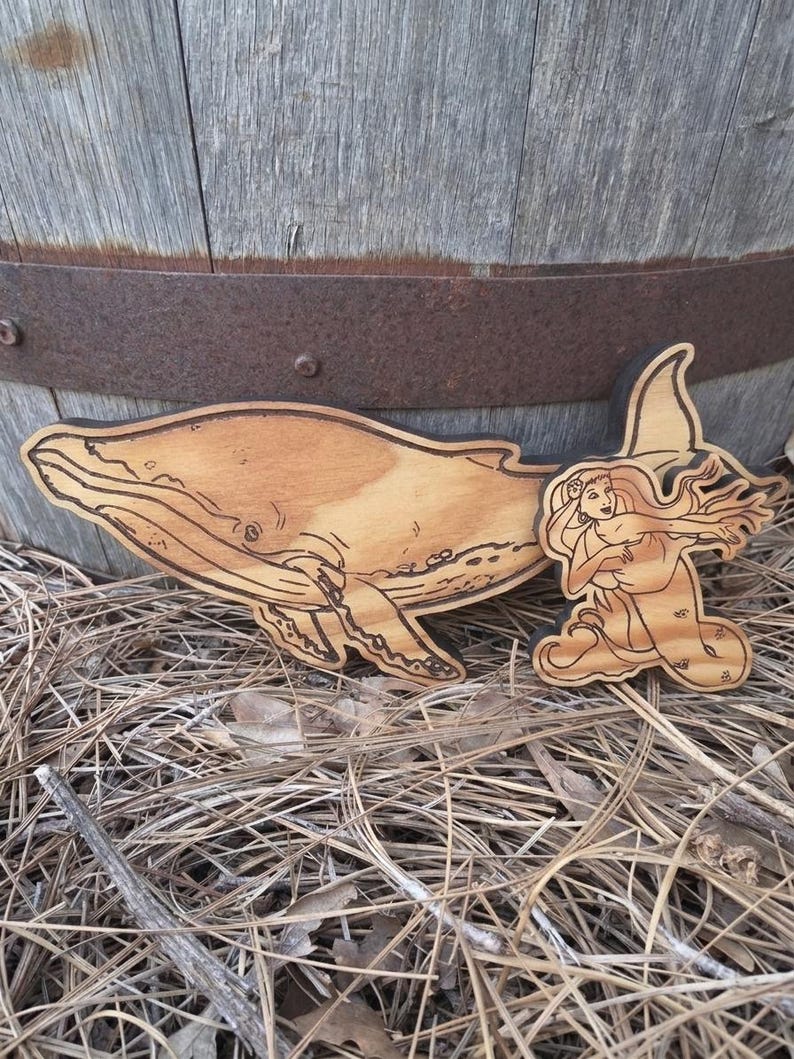 May include: A light brown wooden cutout featuring a detailed engraving of a humpback whale swimming alongside a mermaid with long flowing hair. The wood grain is visible throughout the surface, and the edges of the shapes are dark and defined.