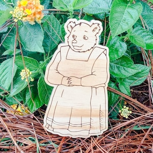 May include: A light-colored wooden cutout featuring a laser-engraved illustration of a bear wearing a long-sleeved dress and an apron. The bear is depicted in a standing position with hands clasped together in front of the torso, showcasing detailed line work.