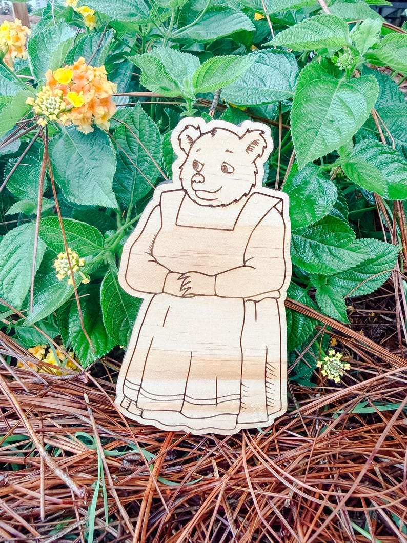 May include: A light-colored wooden cutout featuring a laser-engraved illustration of a bear wearing a long-sleeved dress and an apron. The bear is depicted in a standing position with hands clasped together in front of the torso, showcasing detailed line work.