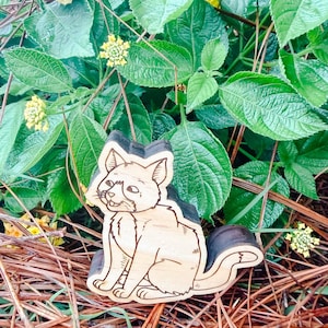 May include: A light-colored wooden cutout of a sitting cat with dark engraved outlines. The cat is depicted in a seated position with its tail curved upward, featuring detailed line work for the fur, ears, whiskers, and facial features.
