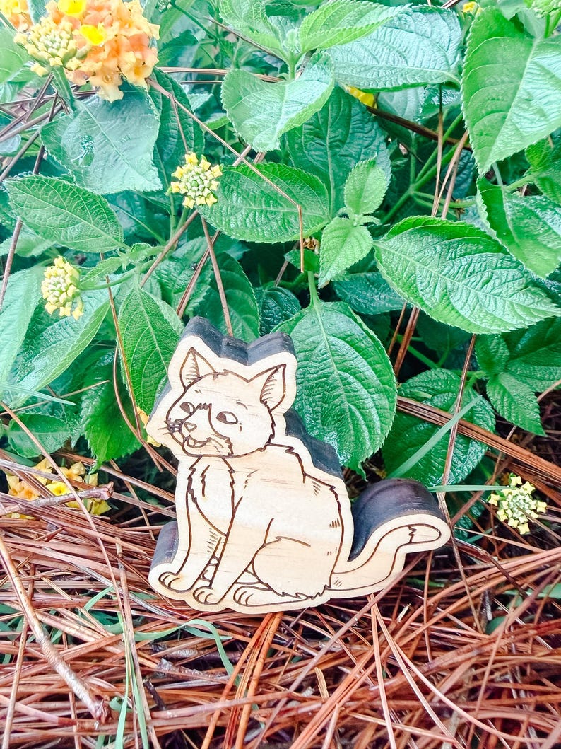 May include: A light-colored wooden cutout of a sitting cat with dark engraved outlines. The cat is depicted in a seated position with its tail curved upward, featuring detailed line work for the fur, ears, whiskers, and facial features.