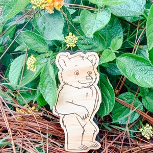 May include: A light-colored wooden cutout shaped like a standing bear with dark engraved outlines defining its facial features, fur texture, and body silhouette. The bear is depicted in a cartoon style with a friendly expression and rounded ears.