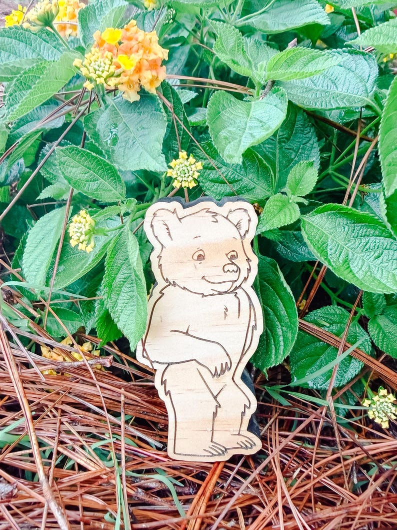 May include: A light-colored wooden cutout shaped like a standing bear with dark engraved outlines defining its facial features, fur texture, and body silhouette. The bear is depicted in a cartoon style with a friendly expression and rounded ears.
