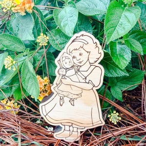 May include: A light-colored wooden cutout featuring a laser-engraved illustration of a person with shoulder-length hair wearing a dress and holding a small doll. The engraving shows clear outlines of the figures, their clothing, and facial features.