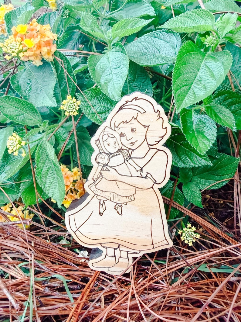 May include: A light-colored wooden cutout featuring a laser-engraved illustration of a person with shoulder-length hair wearing a dress and holding a small doll. The engraving shows clear outlines of the figures, their clothing, and facial features.