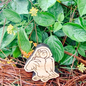 May include: A light-colored wooden cutout shaped like an owl with dark, etched outlines. The owl is depicted in a seated position with its head turned slightly to the side, featuring large eyes and detailed feather markings on its wings and body.