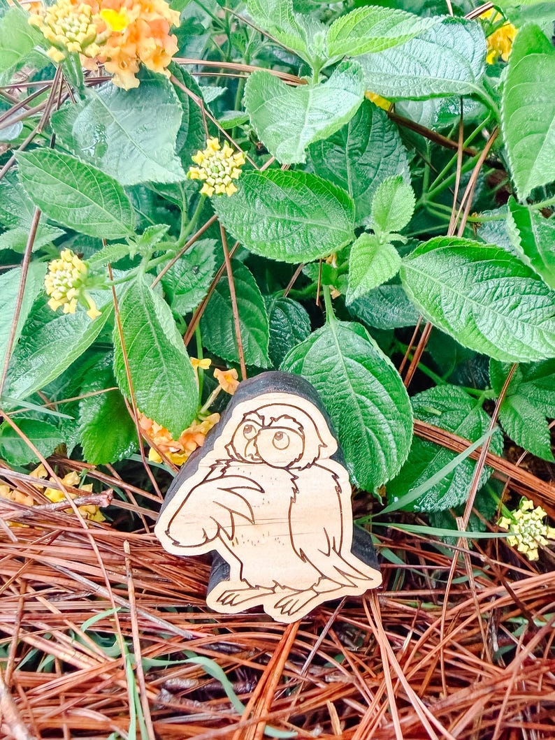 May include: A light-colored wooden cutout shaped like an owl with dark, etched outlines. The owl is depicted in a seated position with its head turned slightly to the side, featuring large eyes and detailed feather markings on its wings and body.