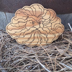 May include: A light brown wooden plaque featuring a laser-engraved illustration of a face with a thick, flowing beard and hair that resembles a cloud. The wood grain is visible throughout the piece, and the engraved lines are dark brown.