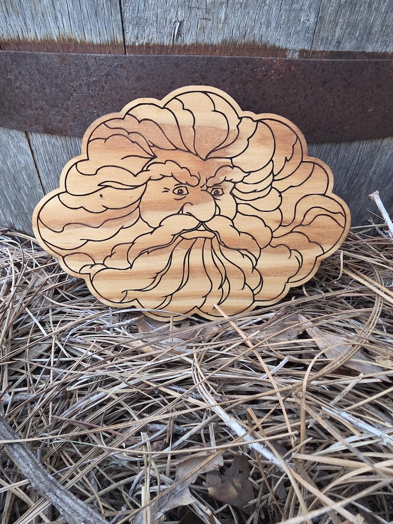 May include: A light brown wooden plaque featuring a laser-engraved illustration of a face with a thick, flowing beard and hair that resembles a cloud. The wood grain is visible throughout the piece, and the engraved lines are dark brown.
