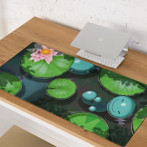 May include: A rectangular desk mat featuring a pond scene with lily pads, a pink water lily, and cartoon-style aquatic creatures. The mat has a dark green and blue colour scheme.