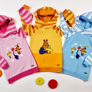 Könnte beinhalten: Drei Kapuzenpullover mit gestreiften Ärmeln und bunten Tierillustrationen. Die Pullover sind rosa, gelb und blau.