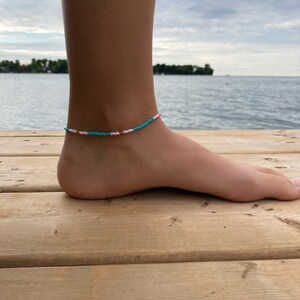 May include: A turquoise and pink beaded anklet with gold accents. The anklet is worn on a person's ankle.