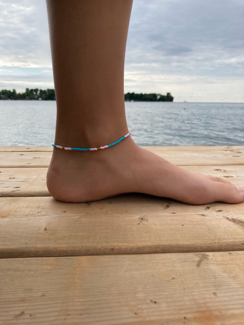 May include: A turquoise and pink beaded anklet with gold accents. The anklet is worn on a person's ankle.