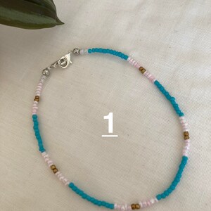 May include: A turquoise, white, and gold beaded anklet with a silver clasp. The anklet is on a white background.