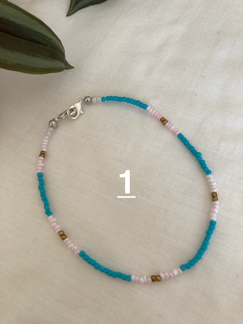 May include: A turquoise, white, and gold beaded anklet with a silver clasp. The anklet is on a white background.