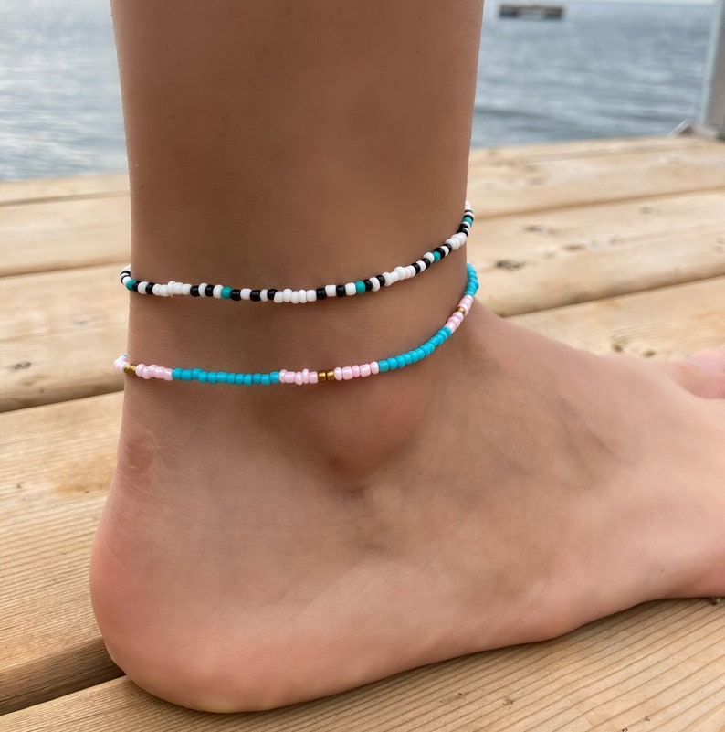 May include: Two beaded anklets on a person's ankle. The top anklet is black, white, and turquoise beads. The bottom anklet is turquoise and pink beads with gold accents.