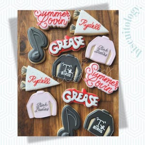 May include: A collection of decorated cookies with a Grease theme. The cookies are shaped like musical notes, jackets with "T Birds" on them, pink sweaters with "Pink Ladies", and the words "Grease", "Summer Lovin'", and "Rydell".