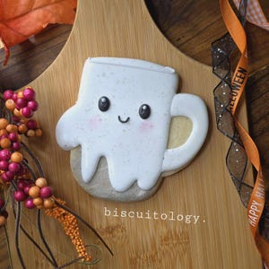 May include: A white and brown decorated sugar cookie shaped like a ghost holding a mug. The ghost has black eyes and a smiling mouth. The cookie is on a wooden surface with the text "biscuitology."