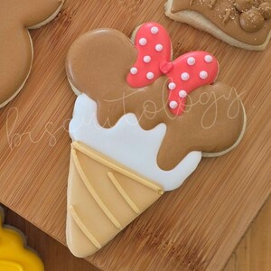 May include: A decorated sugar cookie shaped like an ice cream cone. The cookie features a brown ice cream scoop with a red bow with white polka dots. The cone is tan with yellow lines. The cookie is on a wooden surface.