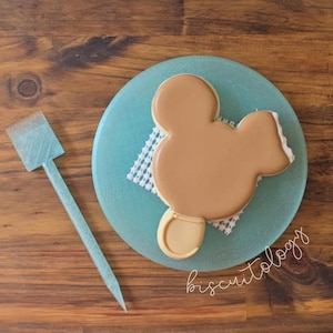 May include: A light brown Mickey Mouse-shaped cookie on a light blue plate with a white doily. A light blue, pointed spatula is to the left. The word "biscuitology" is written in cursive on the plate.