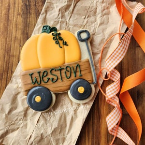 May include: A decorated sugar cookie in the shape of a wagon with a pumpkin on top. The wagon is brown with black wheels and the name Weston is written on the side. The cookie is on a brown paper bag.