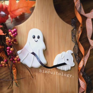 May include: Two Halloween-themed sugar cookies decorated with white icing. A ghost cookie is holding a black leash attached to a small dog cookie. The cookies are on a wooden cutting board with a black and orange ribbon.