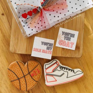 May include: Two heart-shaped cookies decorated with basketball and sneaker designs. The cookies are on a wooden surface with two small cards that read "You're my Sole Mate" and "You're the GOAT".