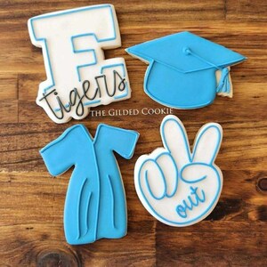May include: Four decorated sugar cookies. One cookie is shaped like a graduation cap, another is shaped like a letter 'E' with the word 'tigers' written below it, another is shaped like a graduation gown, and the last is shaped like a hand making a peace sign with the word 'out' written below it.