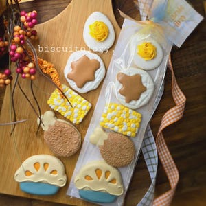 May include: A set of nine decorated sugar cookies in a clear plastic bag. The cookies are shaped like a turkey leg, corn on the cob, a star, a fried egg, a Thanksgiving dinner plate, and a turkey drumstick. The cookies are decorated with brown, yellow, white, and blue icing.