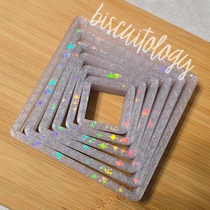 May include: Set of seven silver square cookie cutters with iridescent glitter and rainbow star accents. The cutters are nested inside each other, with the largest cutter at the bottom and the smallest cutter at the top.