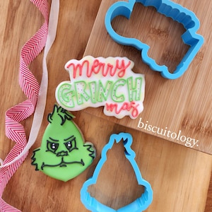 May include: A set of three cookie cutters featuring a Grinch face, a Grinch hat, and the words "Merry Grinchmas". The cutters are blue and are on a wooden surface.