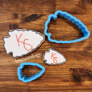 May include: Three cookie cutters in the shape of an arrowhead. Two are blue plastic and one is white with the letters "KC" in orange.