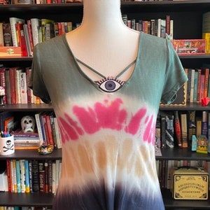 May include: A green, pink, beige, and blue tie-dye shirt with a knotted bottom. The shirt has a criss-cross neckline with a blue eye charm.