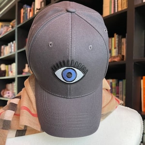 May include: A gray baseball cap with a blue evil eye design embroidered on the front.
