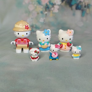 Puede incluir: Una colección de seis figuras de Hello Kitty de varios tamaños. Una figura lleva un sombrero de safari color canela y un chaleco rojo. Otras incluyen figuras con flores azules, un vestido de fresas, un conjunto de invierno rojo y una camisa amarilla.