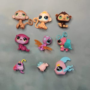 Puede incluir: Una colección de nueve pequeñas figuras de animales de plástico con ojos grandes. El conjunto incluye tres monos en marrón y beige, un gato morado, una criatura rosa con alas de mariposa, un insecto rosa y azul, un rape morado, un pequeño pez globo rosa y un pez rosa.