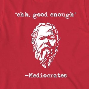 May include: Red t-shirt with a white graphic of a man's face and the text "ehh, good enough" and "Mediocrates".