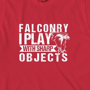 Puede incluir: Camiseta roja con un gráfico blanco que dice "Falconby I Play With Sharp Objects" y una imagen estilizada de un halcón.