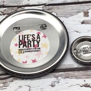 May include: Two metal pinback buttons with the text "LIFE'S A PARTY" in white and pink, surrounded by pink and yellow stars. The larger button is approximately 3 inches in diameter, and the smaller one is about 1 inch.