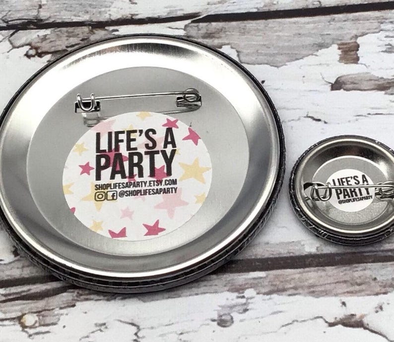 May include: Two metal pinback buttons with the text "LIFE'S A PARTY" in white and pink, surrounded by pink and yellow stars. The larger button is approximately 3 inches in diameter, and the smaller one is about 1 inch.