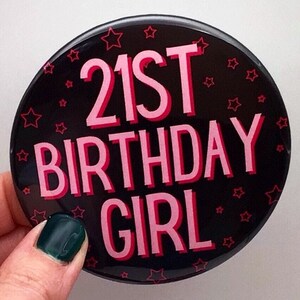 May include: A black button with pink text that reads "21st Birthday Girl" and is decorated with pink stars.