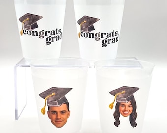 May include: White plastic cups with graduation cap graphics and the words "congrats grad." Two cups feature the graphic and text, while the other two have a graduation cap and a person's face. The cups are on a clear plastic stand.