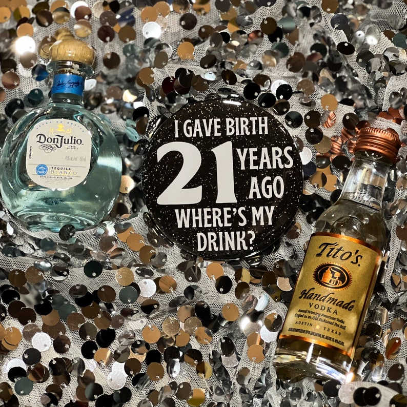 May include: A black button with glitter reads "I gave birth 21 years ago where's my drink?"  Two miniature bottles of alcohol, one clear and one with a white label, are on a silver sparkly background.