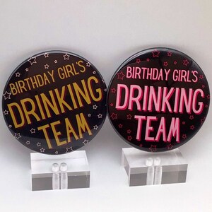 May include: Two black button badges with white stars. One badge says "Birthday Girl's Drinking Team" in gold letters. The other badge says "Birthday Girl's Drinking Team" in pink letters.