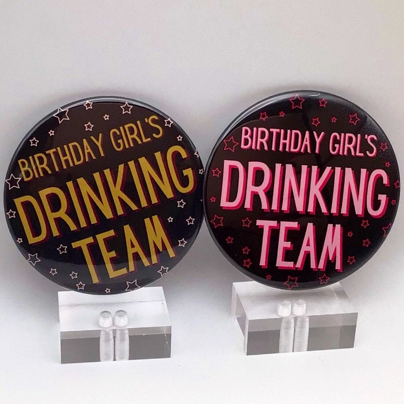 May include: Two black button badges with white stars. One badge says "Birthday Girl's Drinking Team" in gold letters. The other badge says "Birthday Girl's Drinking Team" in pink letters.