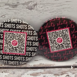 May include: Two black and white round buttons with a pink and white QR code and the text "shots" repeated in pink. The QR code has a pink icon of a cocktail glass.